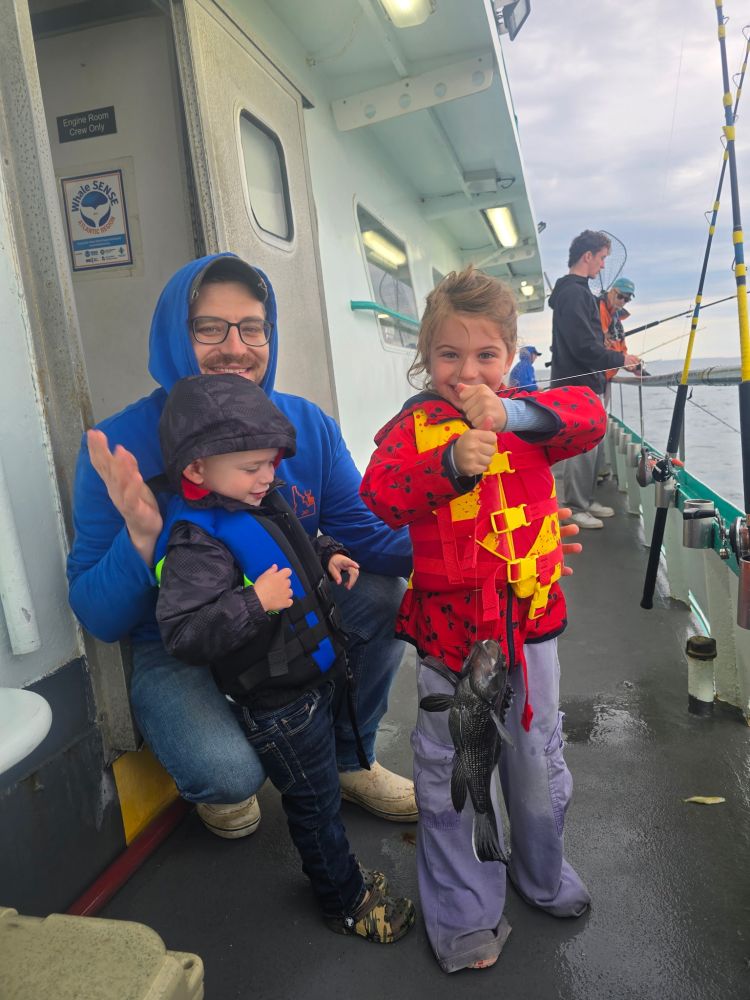 Two kids in life jackets on boat, one holding a fish, with an adult smiling beside them.