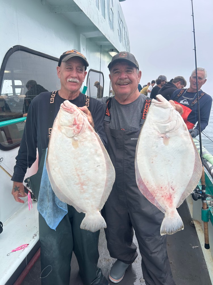 Two men on a boat holding large flat fish, smiling at the camera.