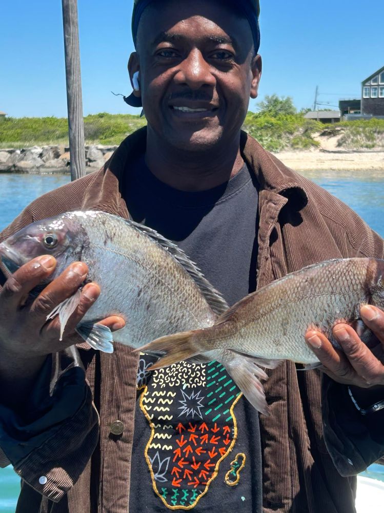 Person holding two fish with water and greenery in the background.