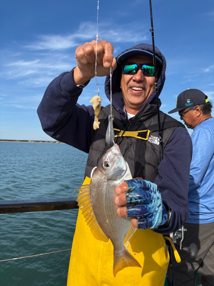 Man in yellow overalls holding a fish on a fishing line, standing on a boat.