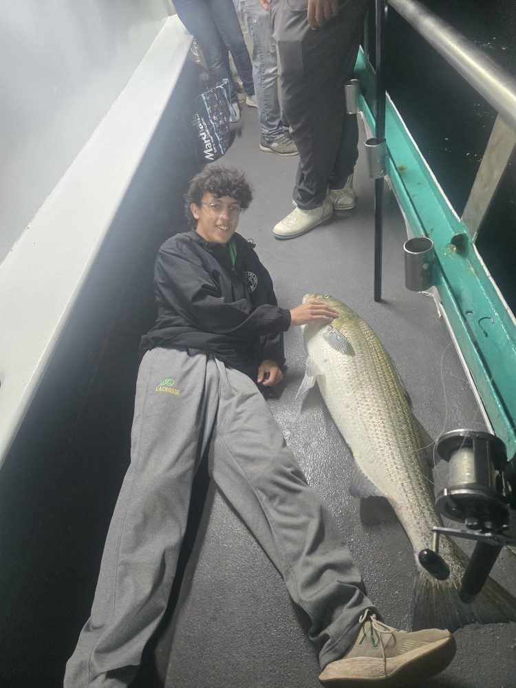 Person lying on a boat deck holding a large fish, with fishing gear beside.