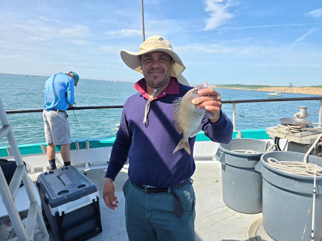 Man holding a fish on a boat, wearing a hat and blue sweater.