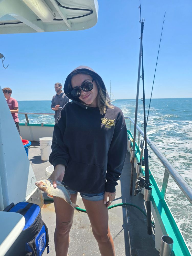 Person in hoodie holding a small fish on a boat with fishing rods, ocean in background.