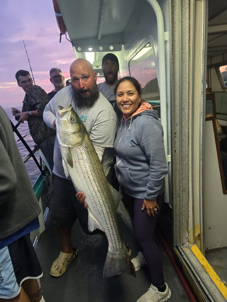 Group on a boat holding a large fish, smiling at sunset.
