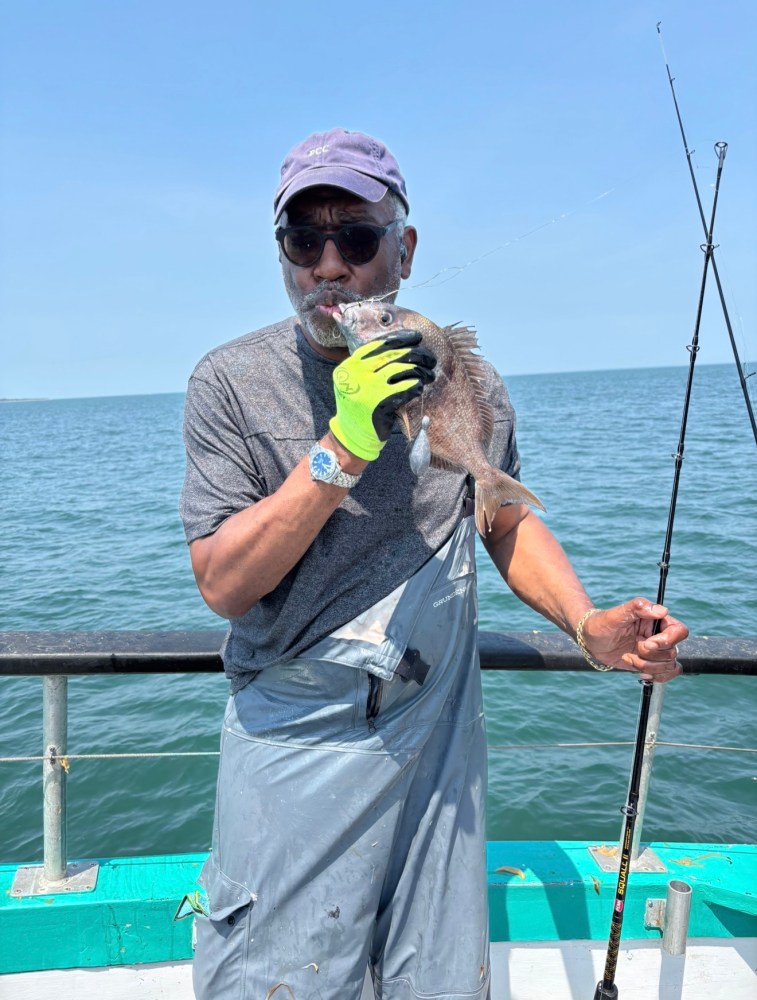 Person in fishing gear holding a fish on a boat, with ocean in the background.