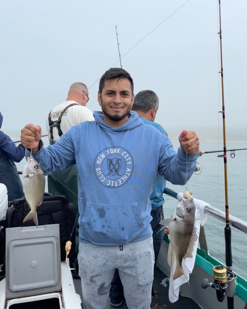 Man on boat holding two fish, wearing blue hoodie, with others in background.