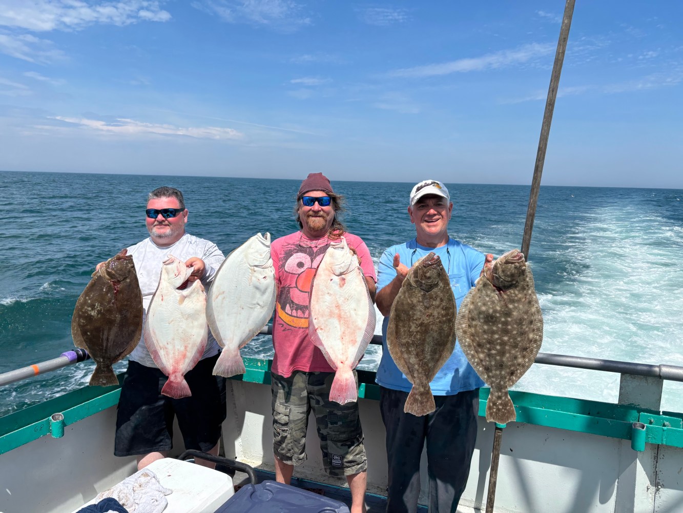 Three people on a boat holding large flatfish by the ocean.