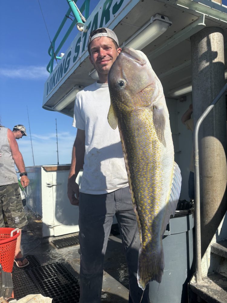 Man holding a large fish on a boat deck under a blue sky.
