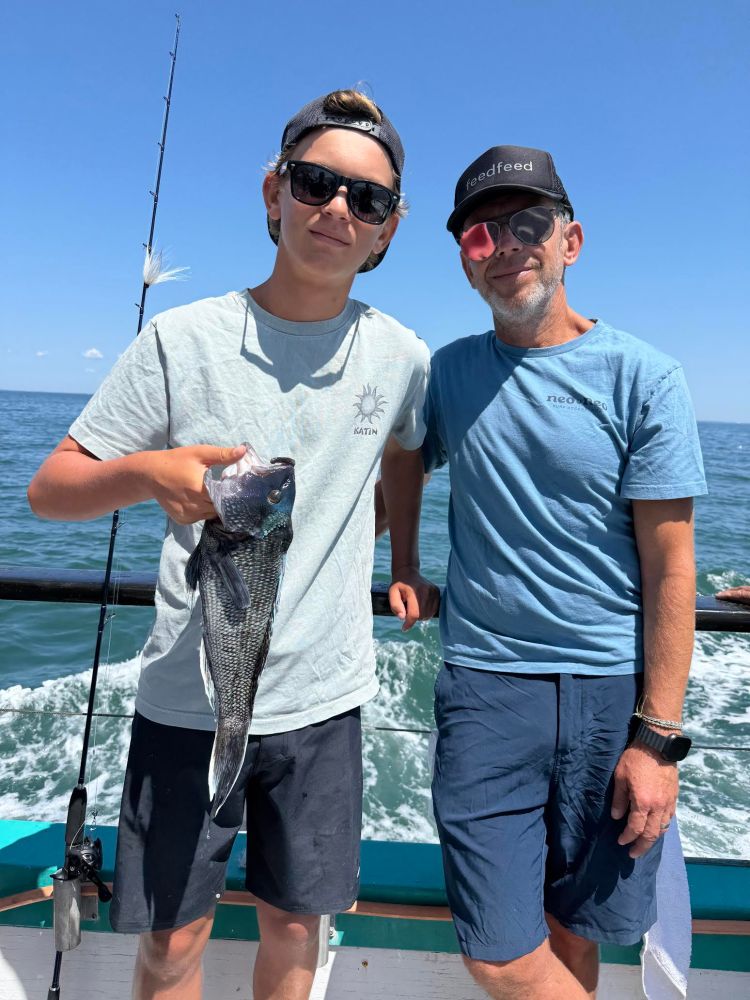Two people stand on a boat, one holding a fish, with ocean in the background.