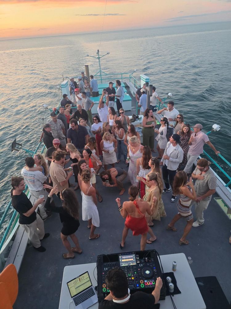 People dancing on a boat deck at sunset with a DJ playing music.