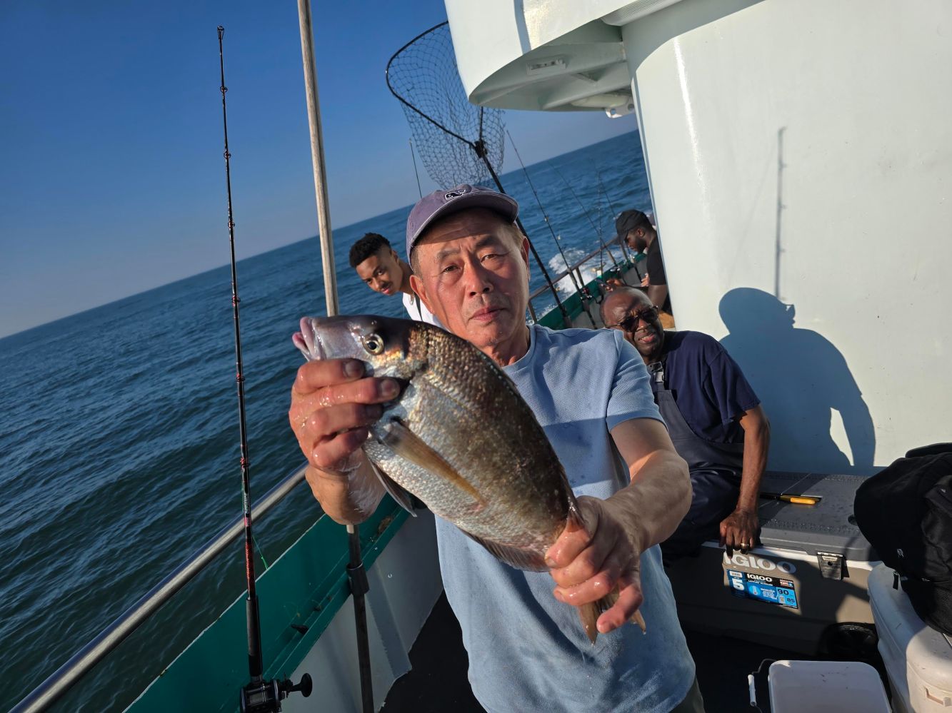 Man on boat proudly holds up a caught fish, with others sitting nearby.