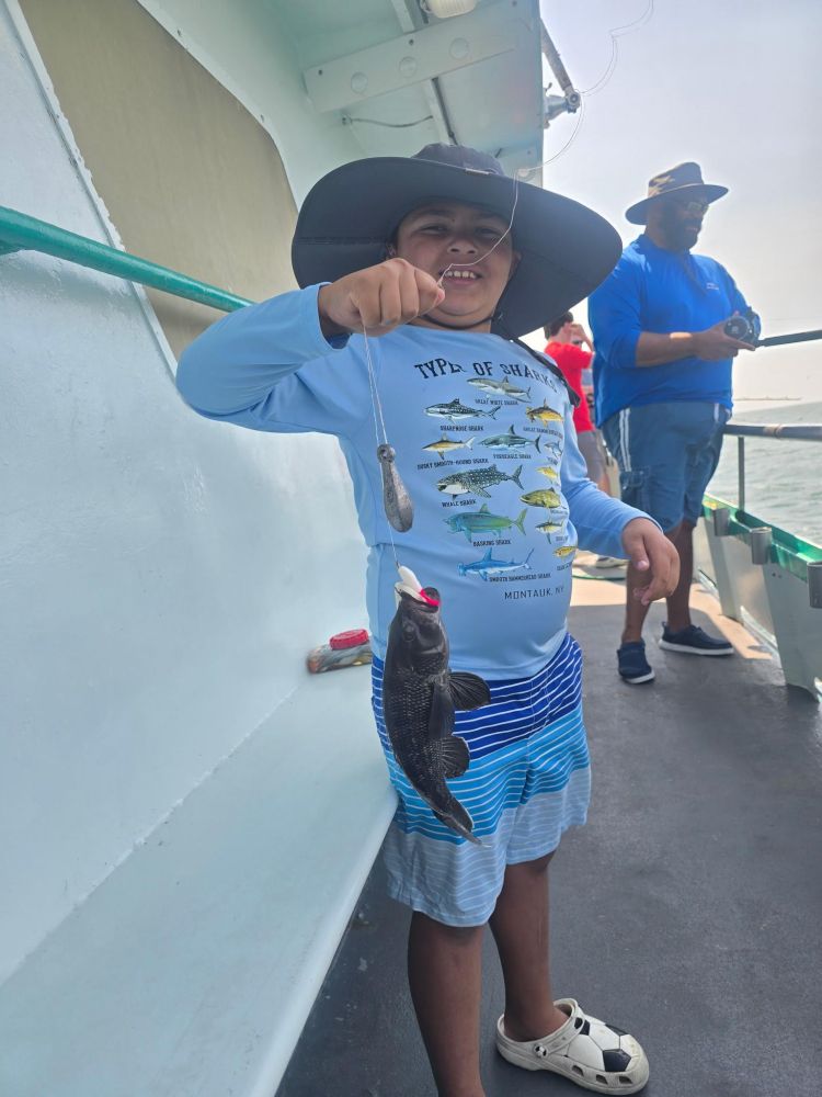 Child on boat holding fish with fishing line, wearing a large hat and a fishing-themed shirt.