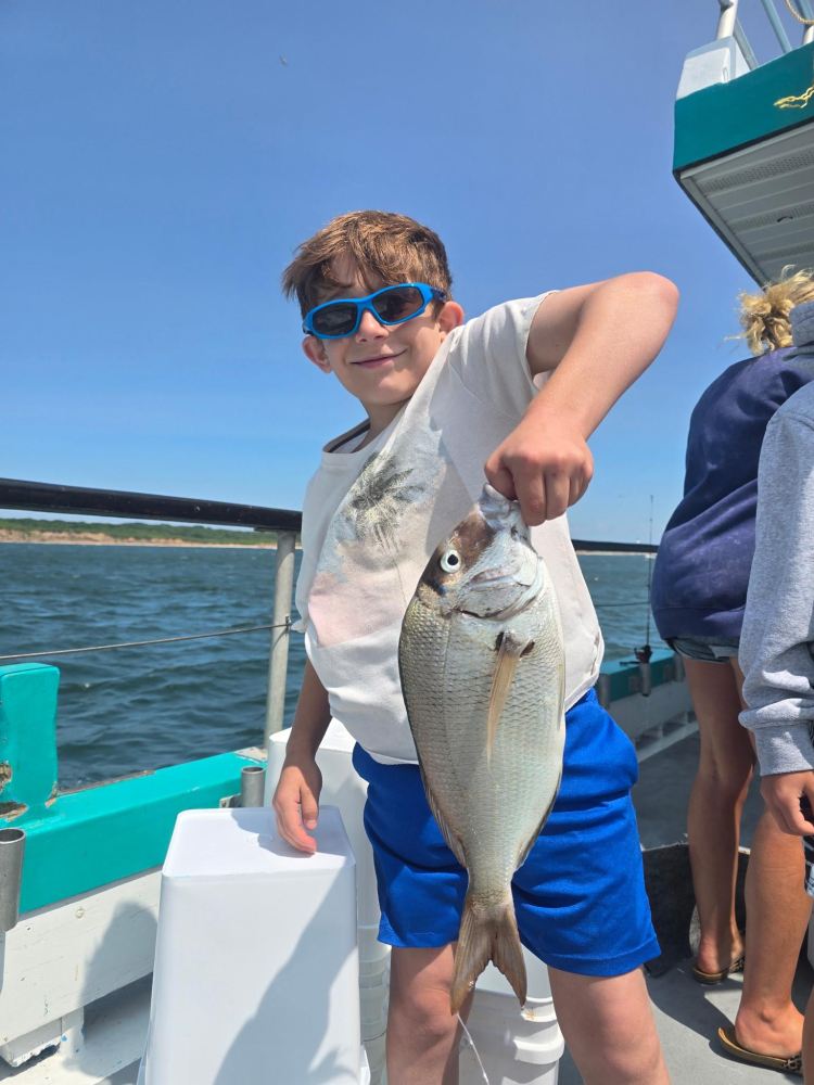 Boy on boat holding a fish, wearing blue sunglasses and shorts.