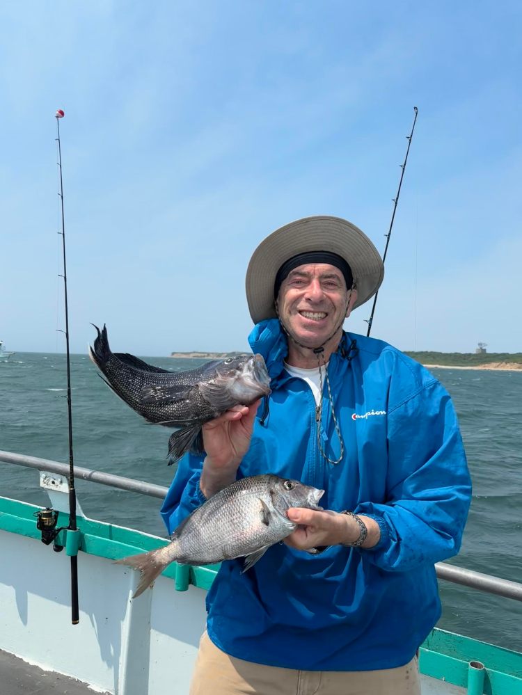Person smiling on a boat holding two fish, ocean in the background.