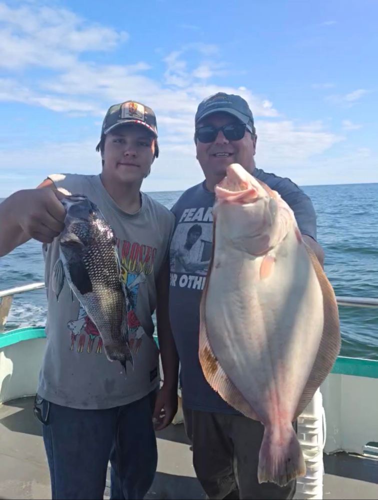 Two people on a boat holding fish, one a flounder and the other a black sea bass.