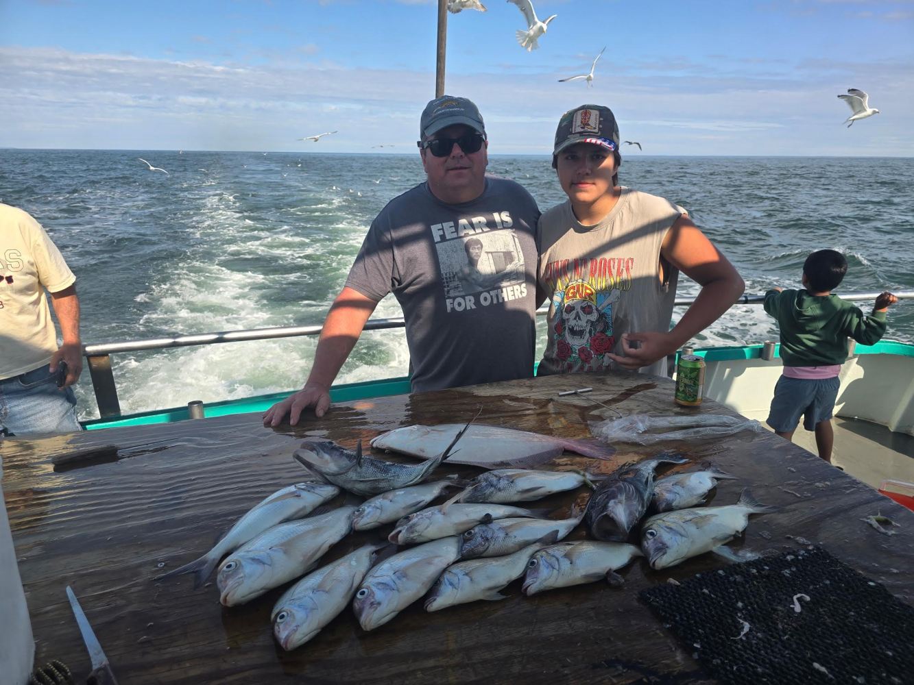 Two men on a boat with caught fish on a table, seagulls flying overhead.