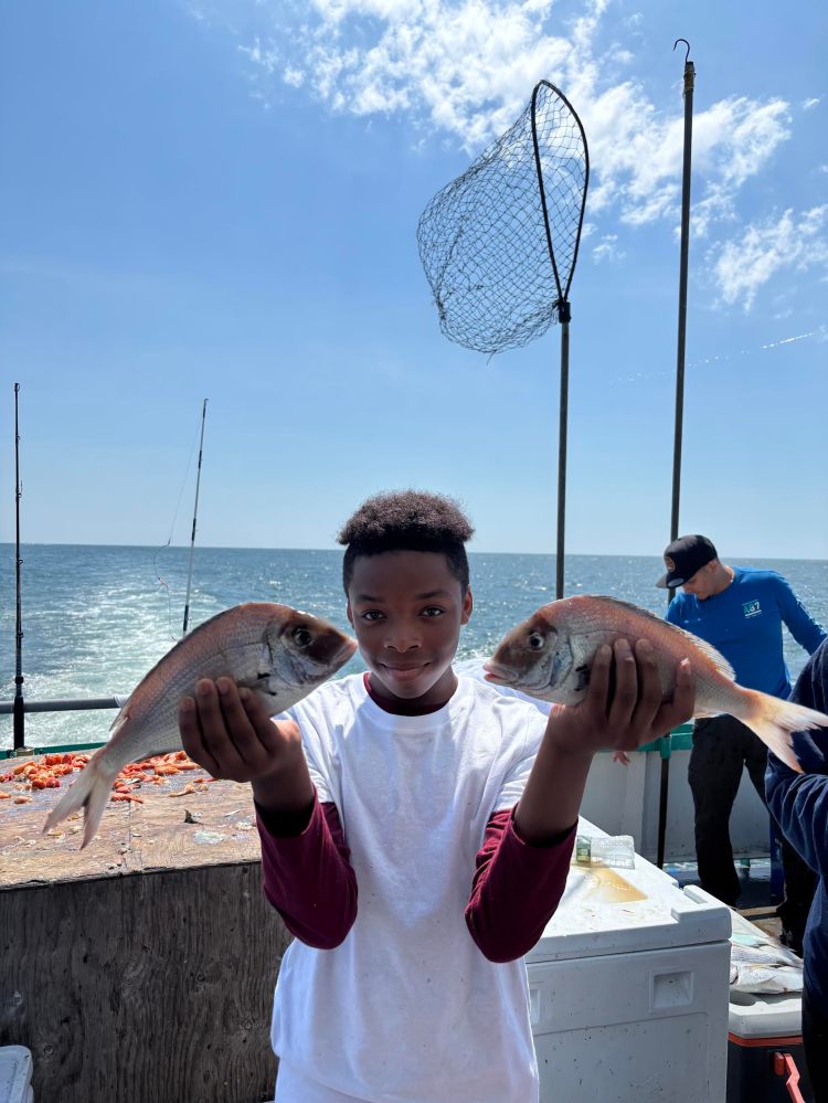Boy holding two fish on a boat with fishing net and ocean in background.