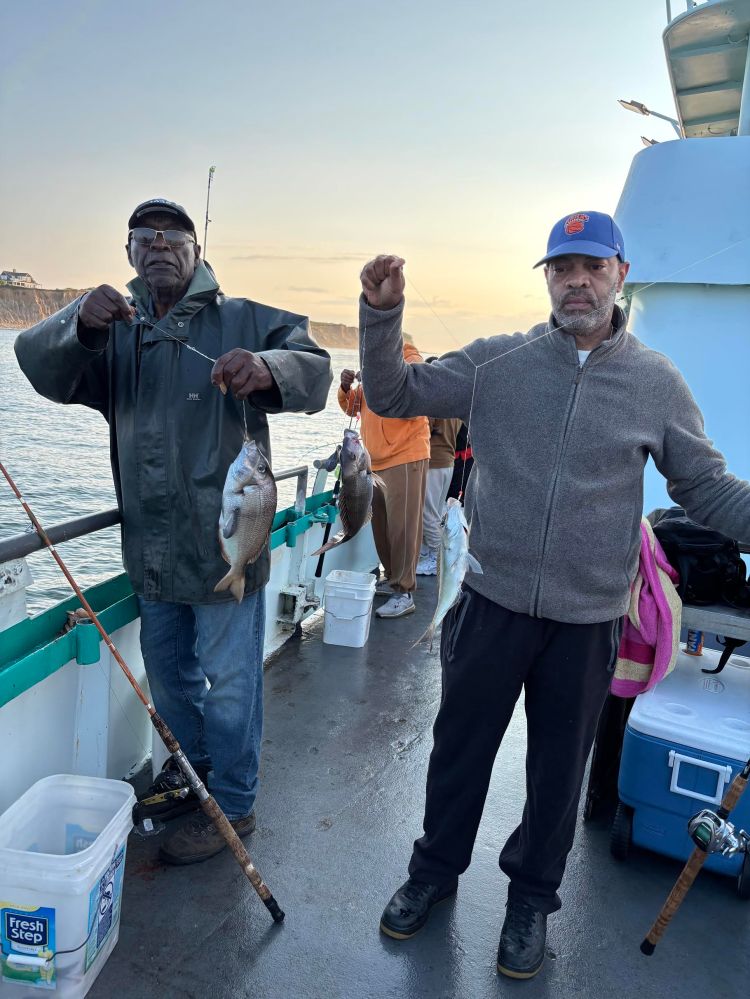 Two men on a boat holding up caught fish at sunset.
