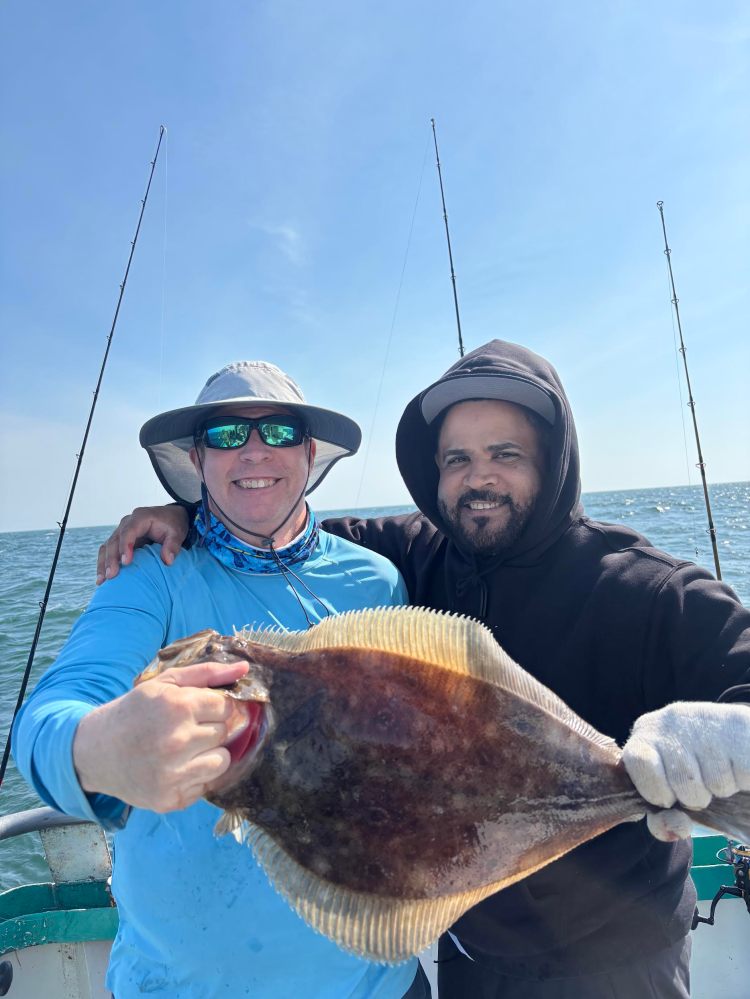 Two people on a boat holding a large fish, smiling. Clear sky and sea in the background.