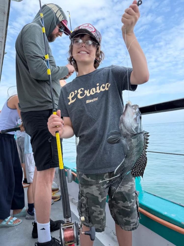 Boy holding a fish on a boat, wearing a cap, next to a man with a fishing rod.