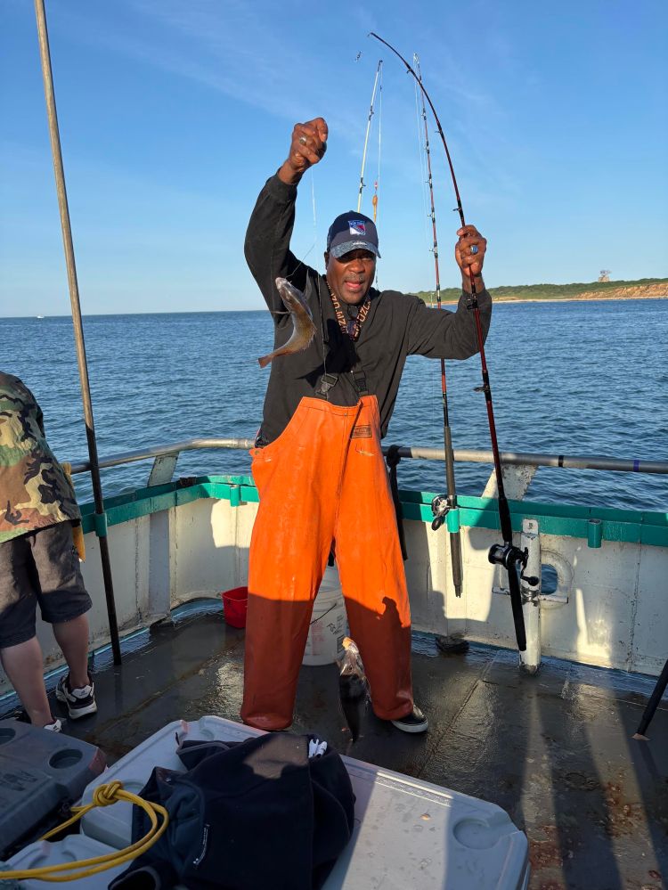 Person on boat holding fish and fishing pole, wearing orange overalls.