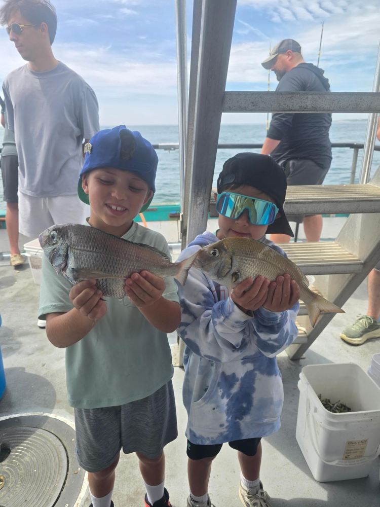 Two kids on a boat holding fish, wearing caps and sunglasses.