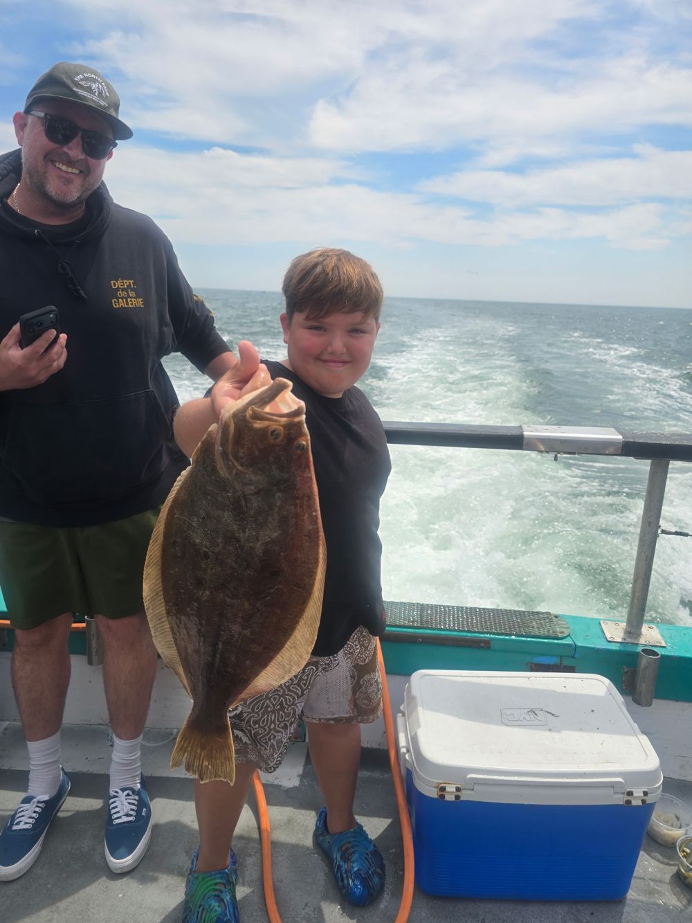 Child proudly holding large fish on a boat with smiling adult and ocean view.