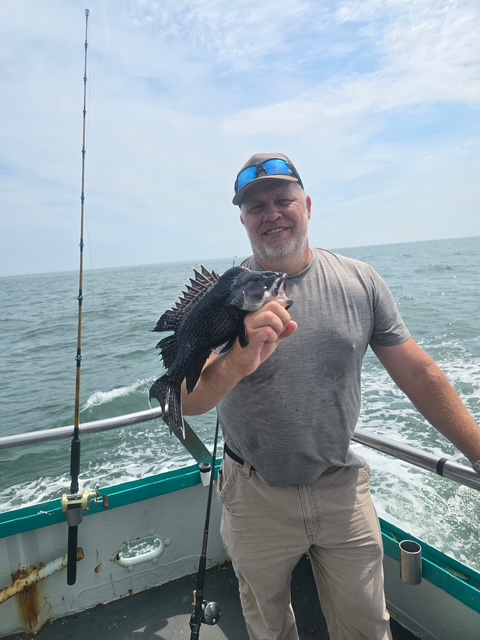 Man on boat holding a fish next to fishing rod with ocean in background.