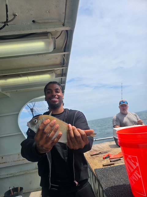 Person on boat holding a fish, another person in the background, ocean and sky visible.