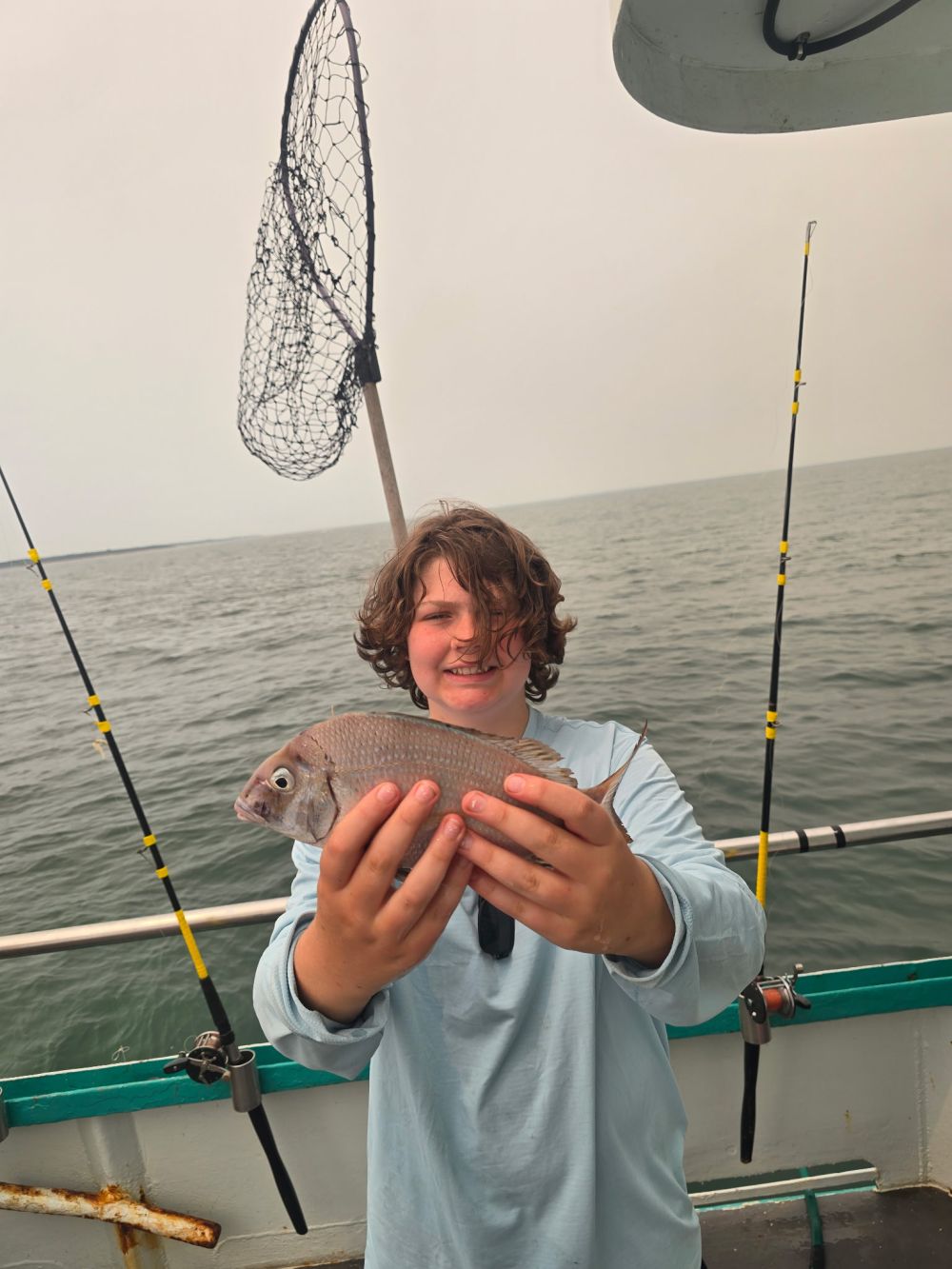 Person on a boat smiling and holding a fish with a net and fishing rods visible.