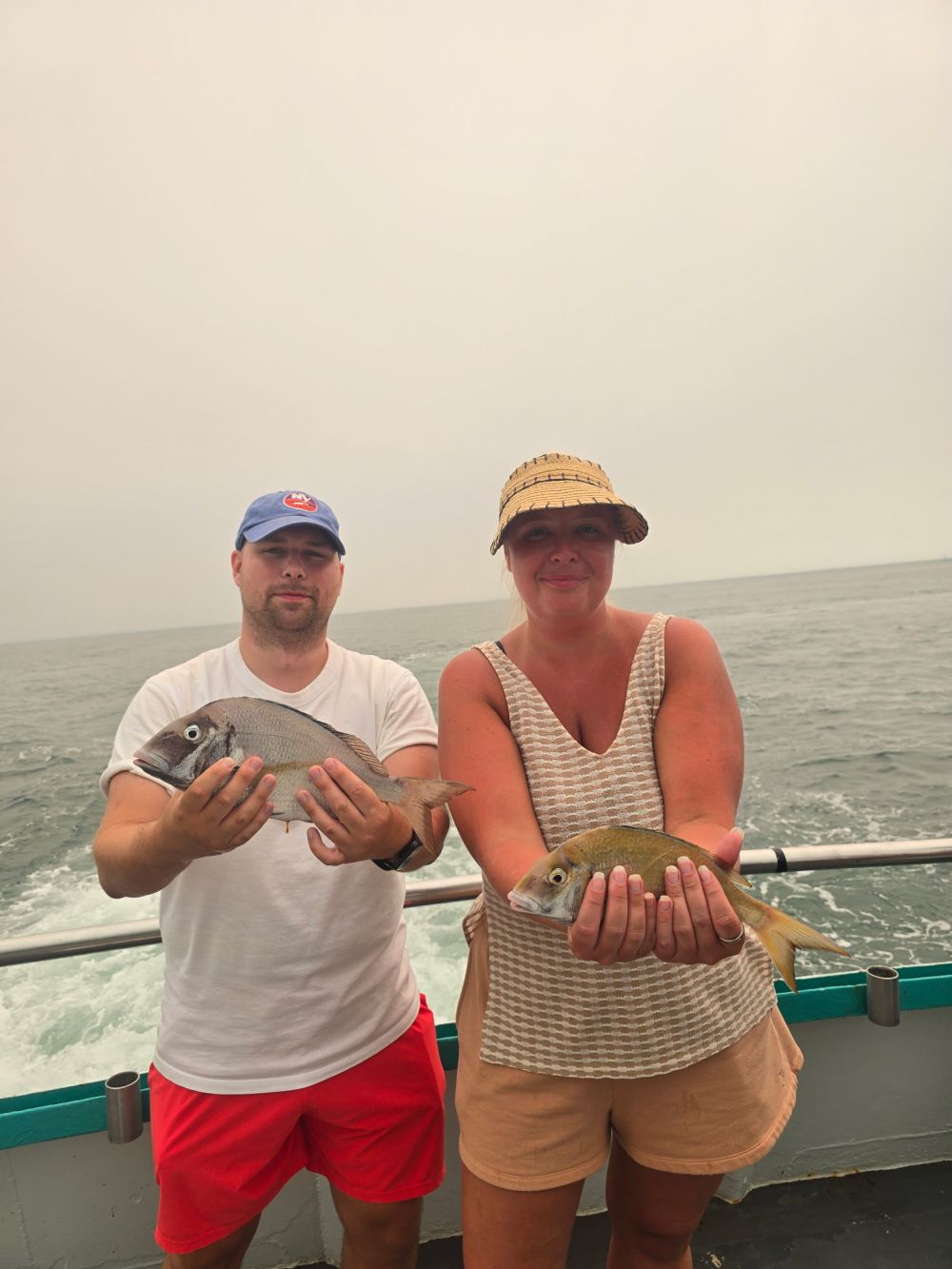 Two people on a boat holding fish near the ocean.