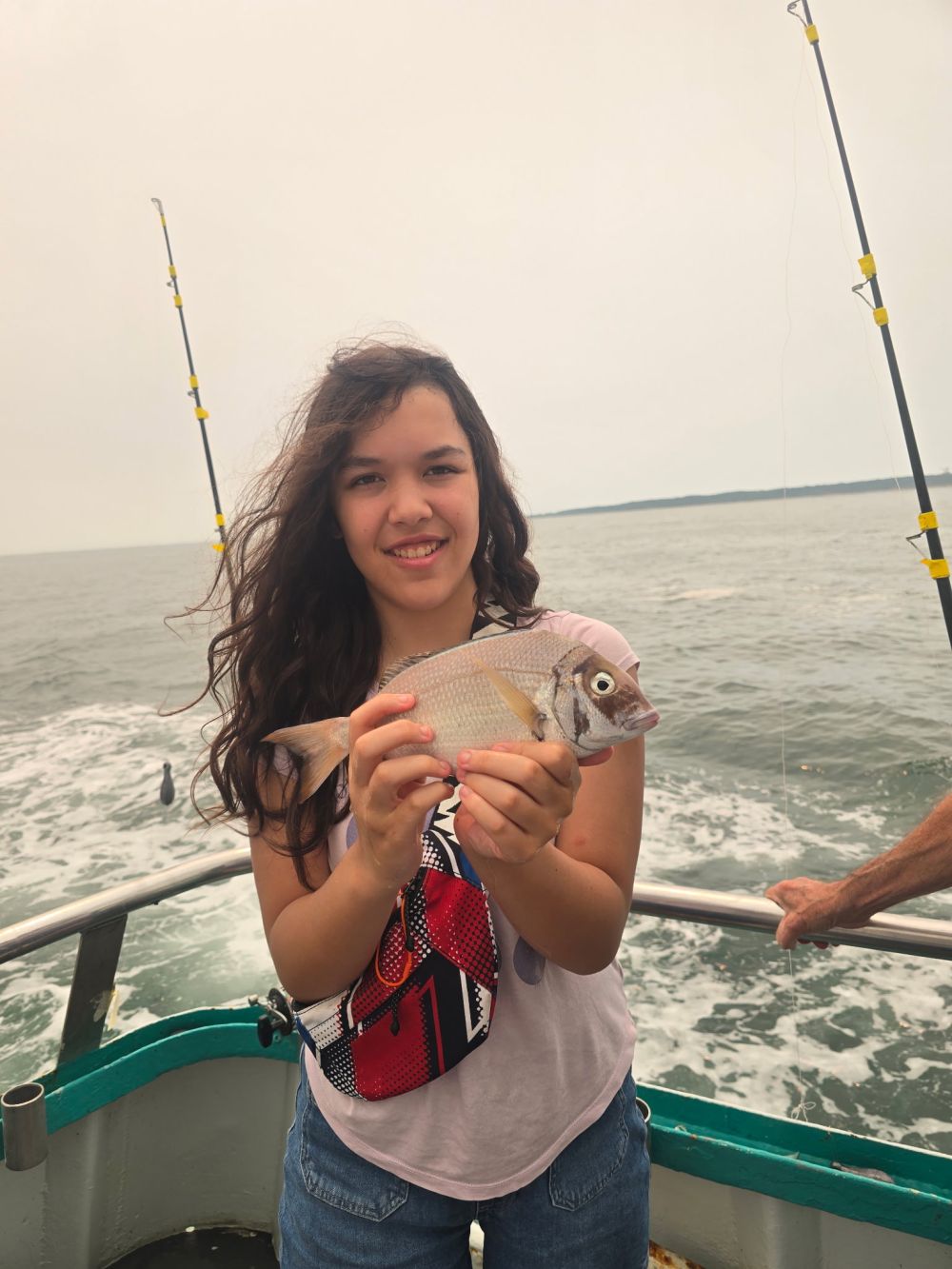 Person holding a fish on a boat with two fishing rods, ocean in the background.