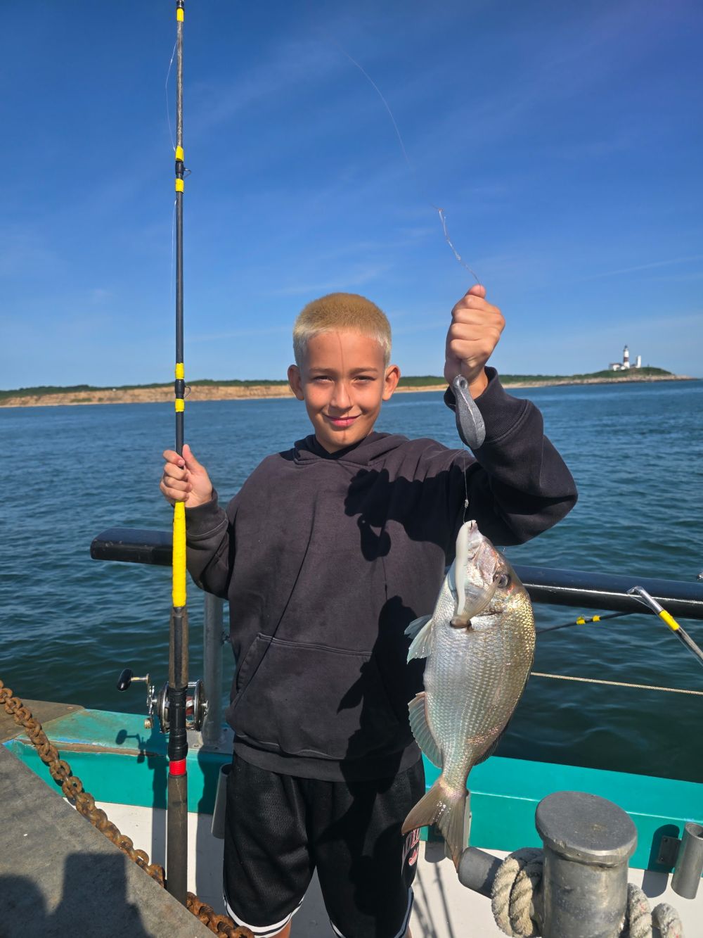 Boy with fishing pole holding a fish on a boat under clear blue sky.