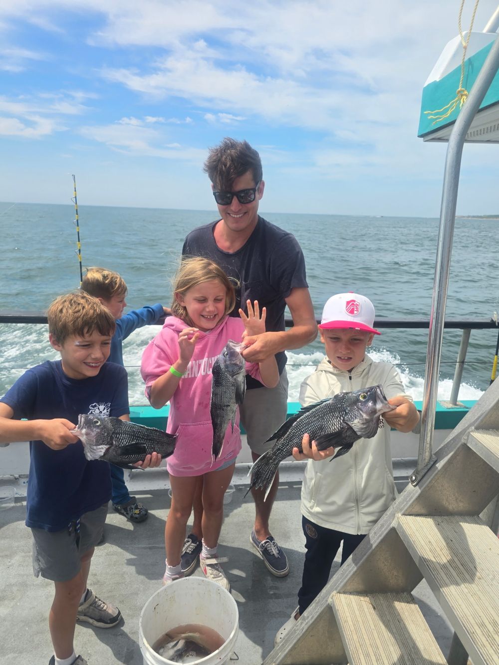 Four children and an adult holding fish on a boat with ocean background.