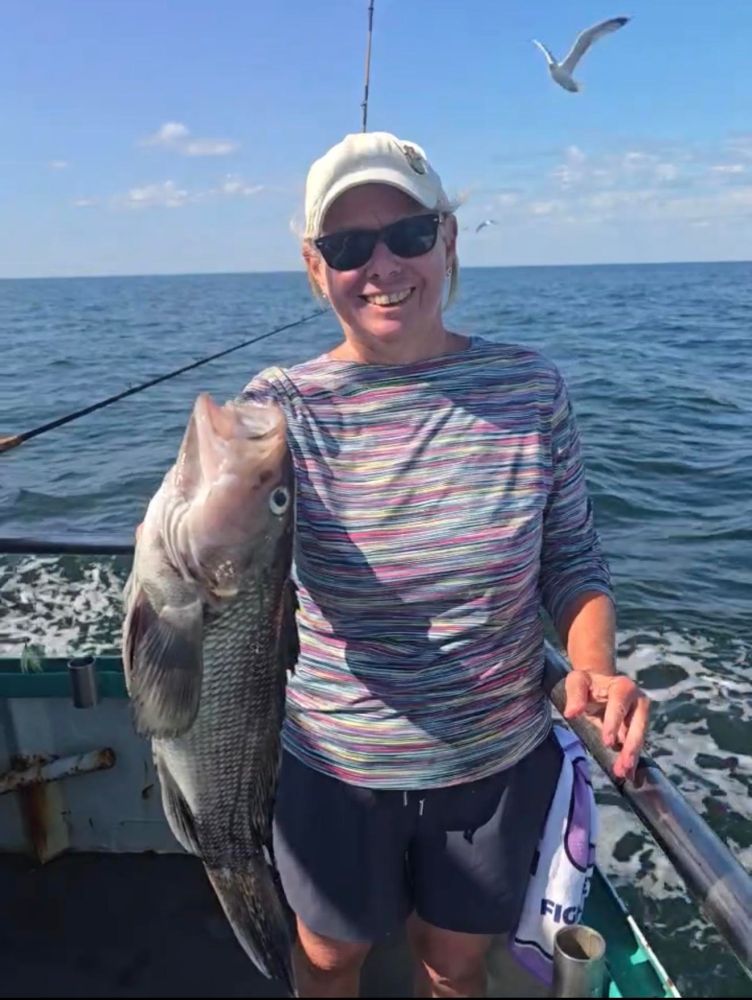 Smiling person holding a large fish on a boat at sea, with a seagull flying overhead.