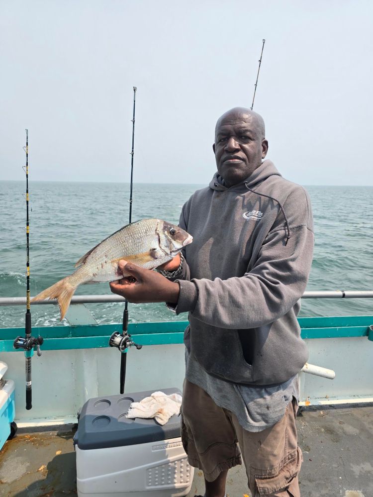 Man holding a fish on a boat with fishing rods and ocean background.
