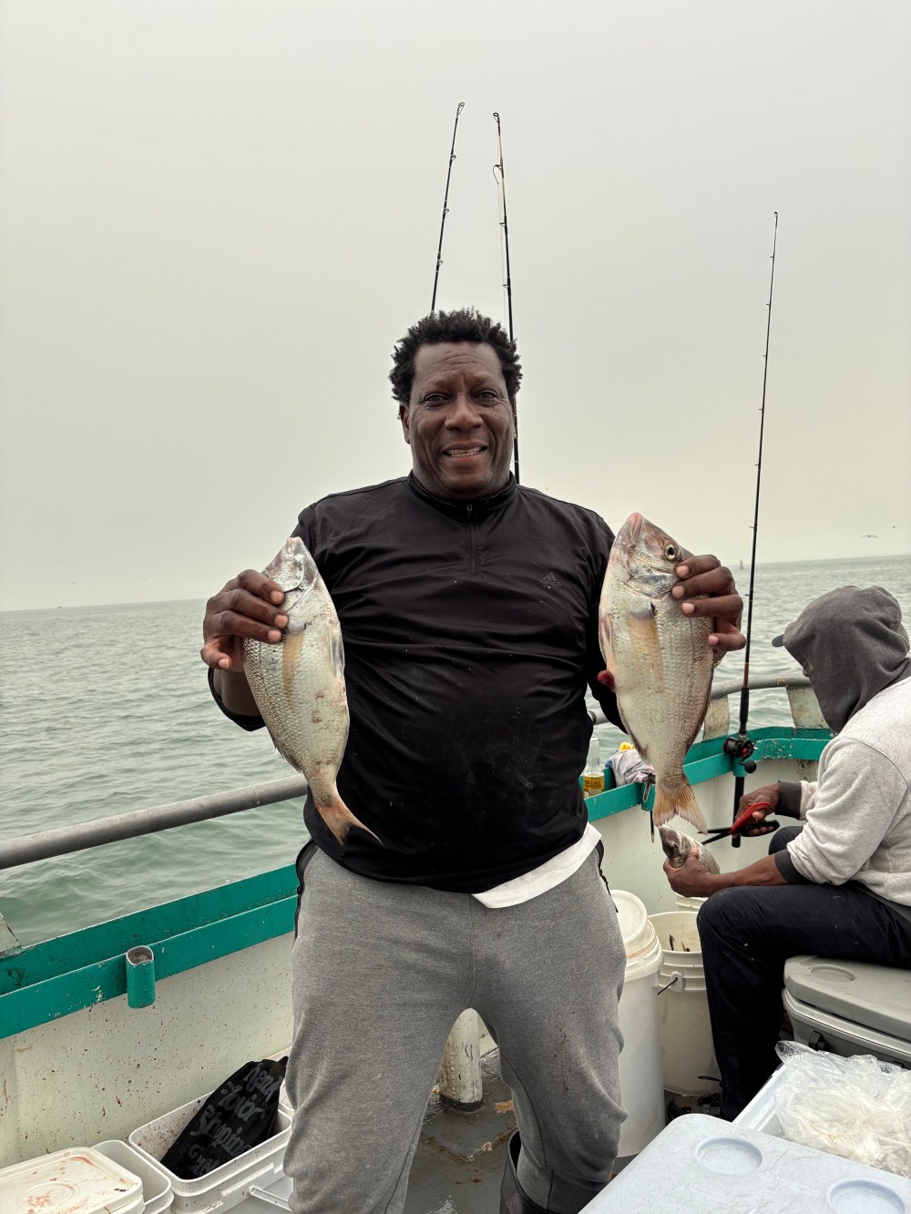 Man smiling holding two fish on a boat with fishing rods in the background.