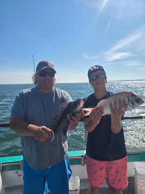 Two people on a boat holding fish, with the ocean in the background.