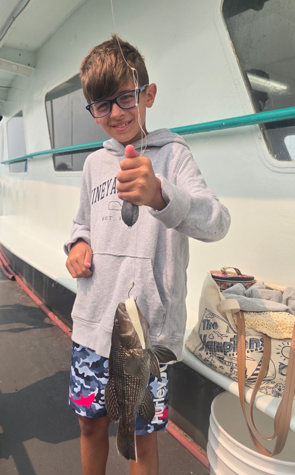 Boy holding a fish on a boat, smiling, wearing glasses and casual clothing.