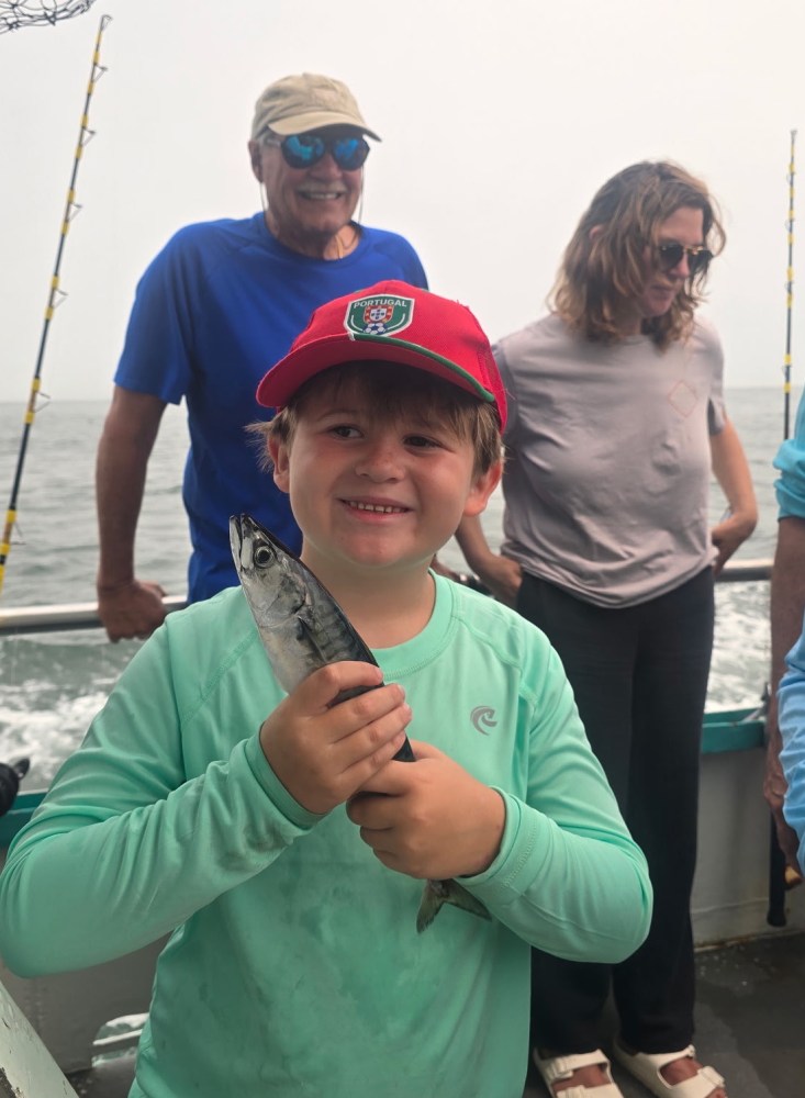 Boy on boat holding a fish, smiling, with two adults in the background.