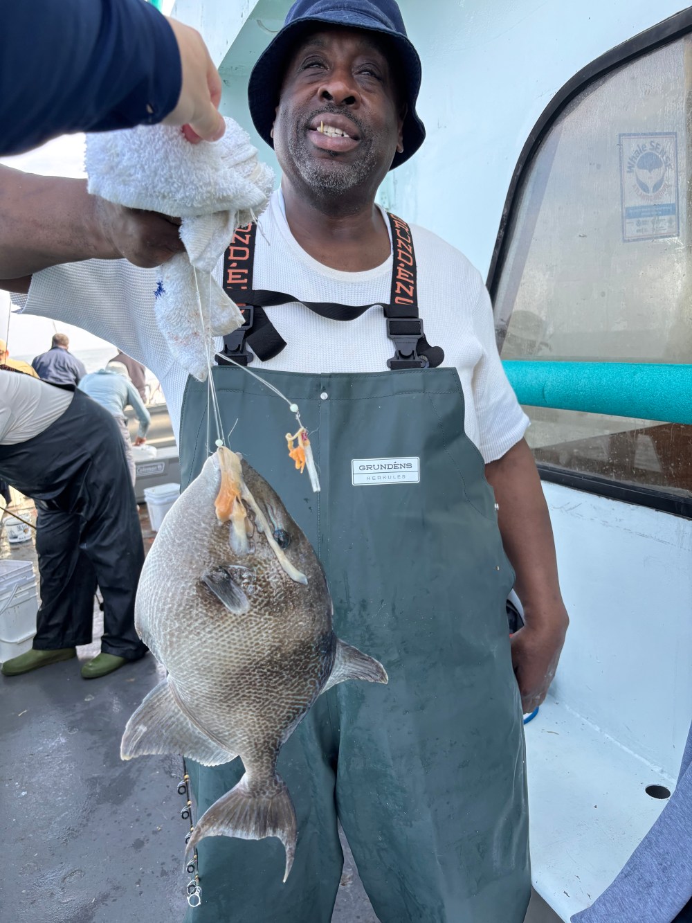 Man holding a large fish with a fishing line on a boat deck, wearing overalls and a hat, smiling.