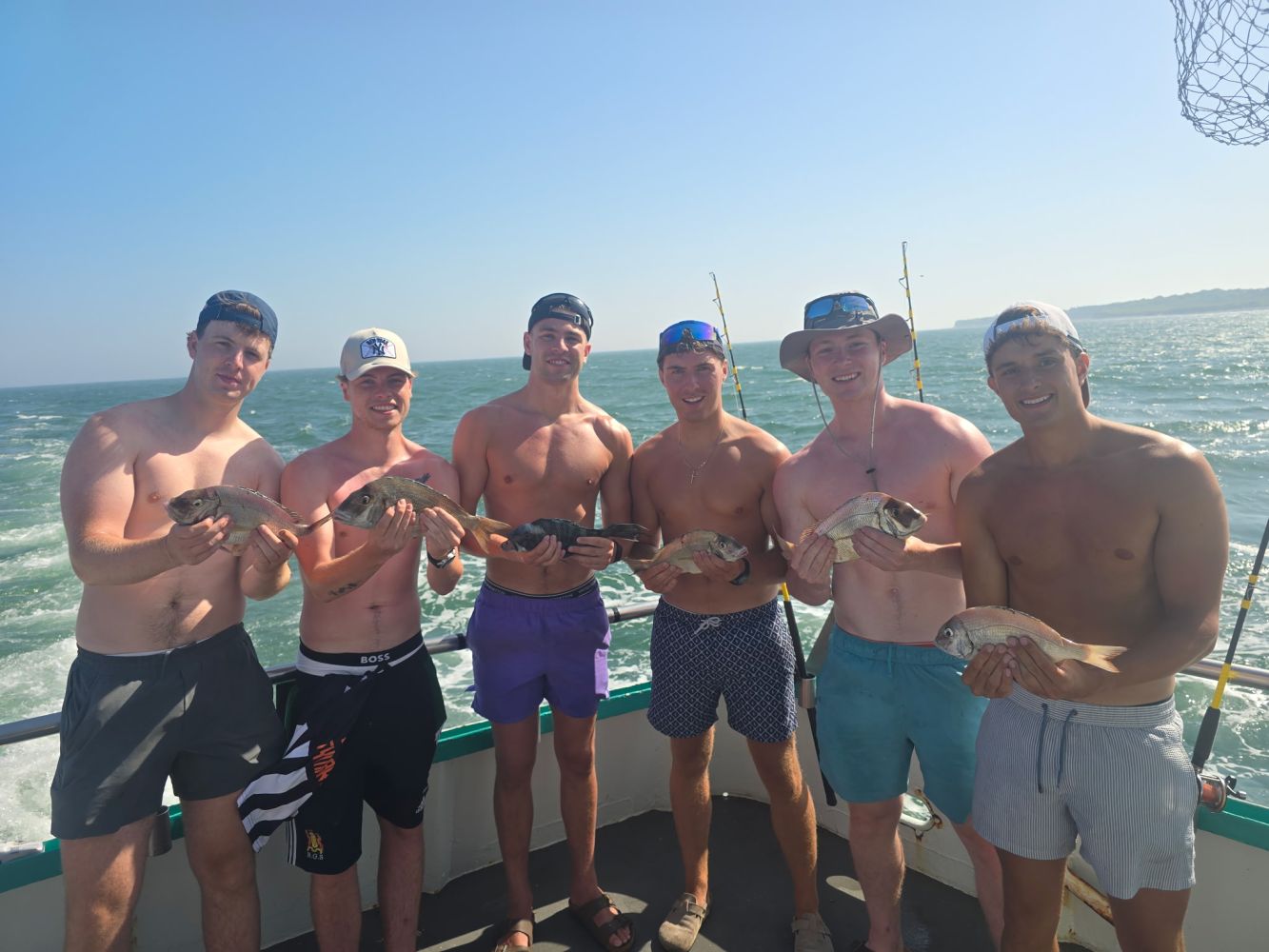 Six men on a boat holding fish with the ocean in the background.