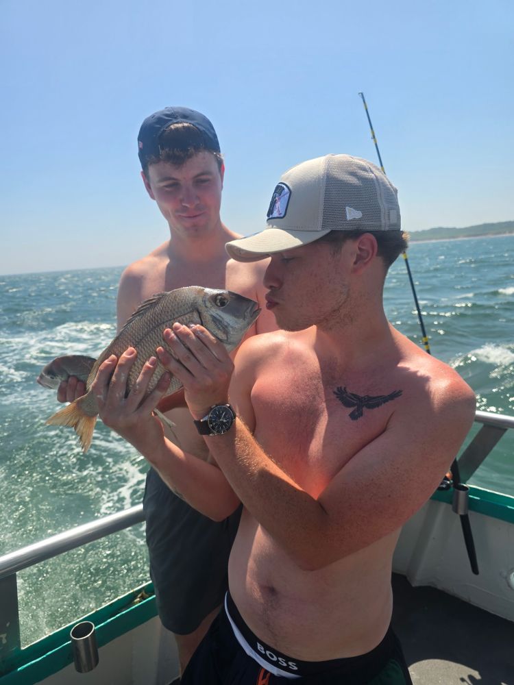 Two shirtless men on a boat, one kissing a caught fish.