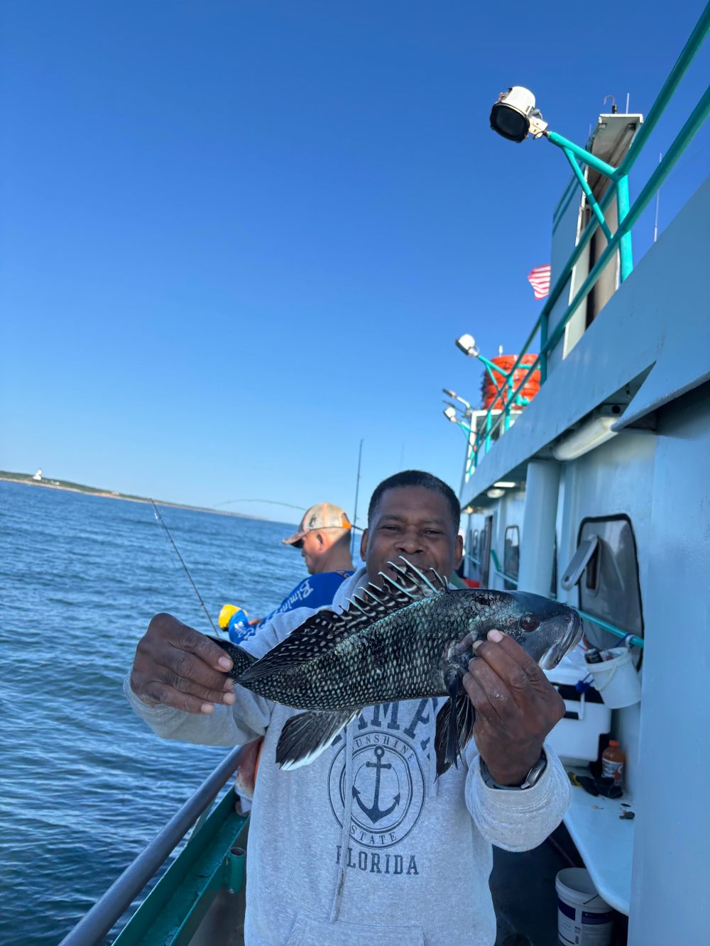 Man smiling, holding a fish on a boat with clear blue sky in the background.