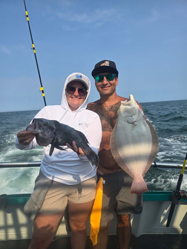 Two people smiling on a boat holding fish, one holds a black fish and the other a flat fish.