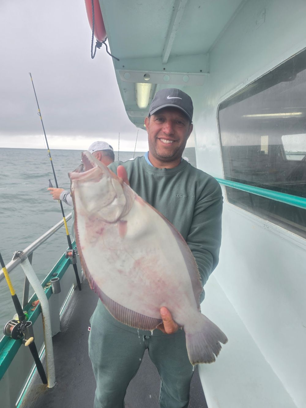 Man on boat holding a large flat fish, with another person fishing in the background.