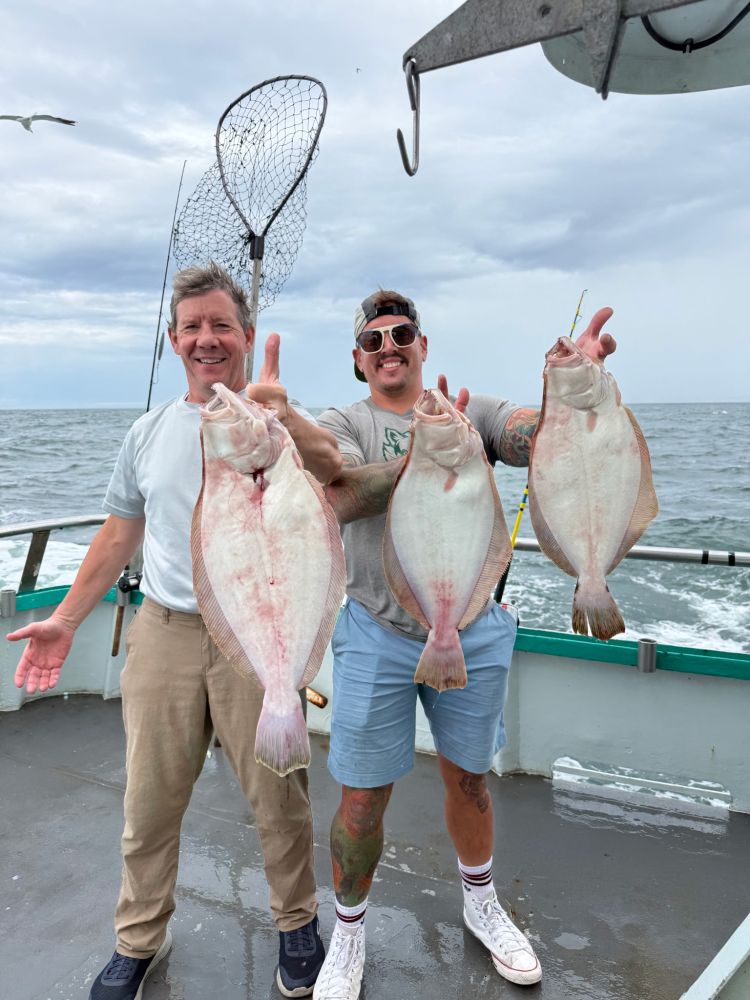 Two men on a boat holding three large fish, with ocean background.