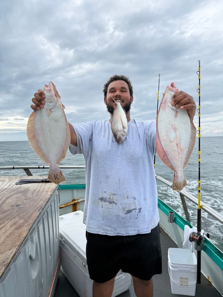 Man on boat holding two flat fish with one in mouth, cloudy sky.