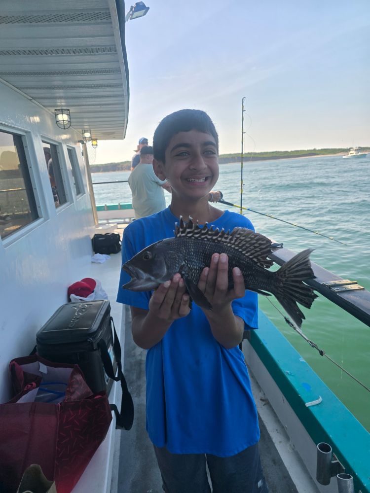 Smiling boy in blue shirt holding a fish on a boat with water in the background.