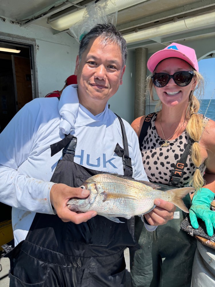 Two people on a boat, one holds a fish; both smiling, wearing casual fishing attire.